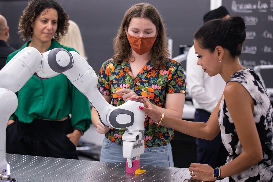Three women stand around a white robotic arm as it picks up a pink cube.